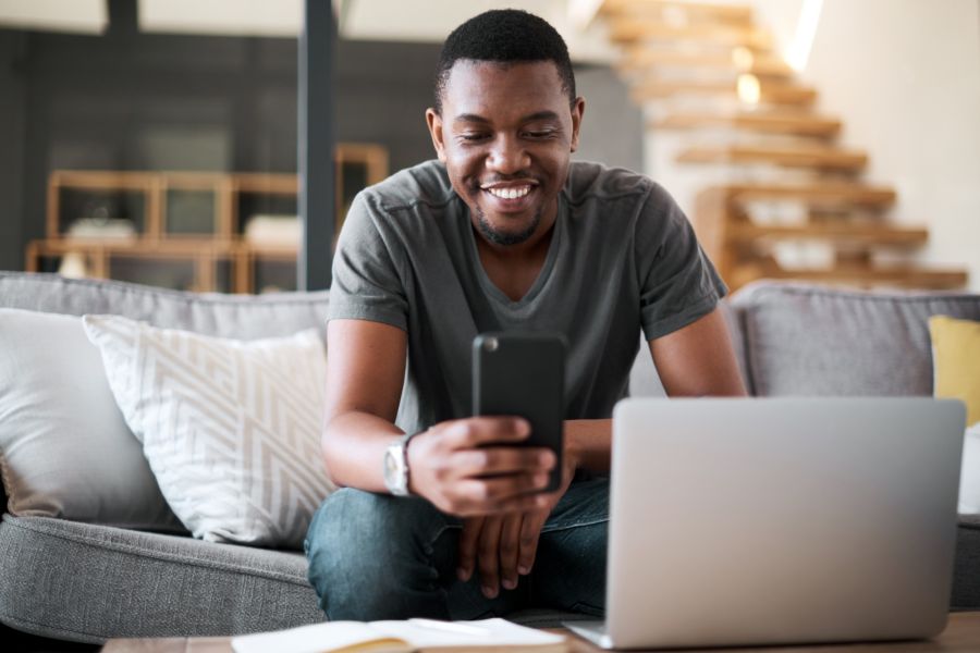 Smiling man on a couch checking his phone beside a laptop in a living room