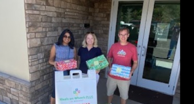 Three people stand outside a building holding wrapped gifts beside a Meals on Wheels Plus donation bin